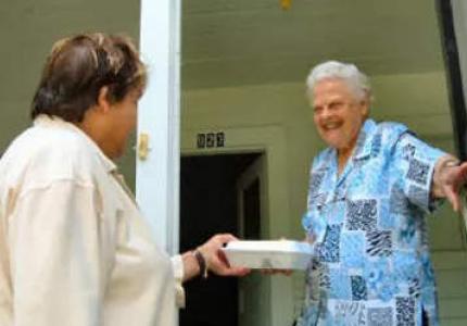 Woman handing a box of food to a senior woman who is holding the door open.