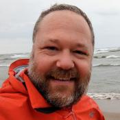 Man in orange jacket standing on beach with body of water in the background.