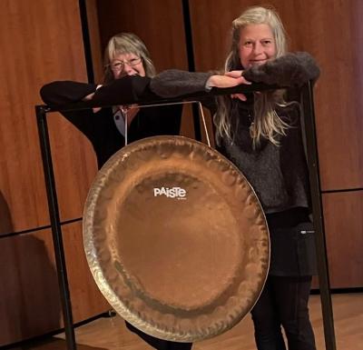 Two women standing behind a big hanging gong-like object.