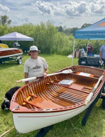 Smiling man in fishing-type hat standing next to a beautifully restored rowboat. Cute dog in background.