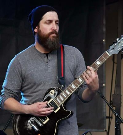 Bearded man in black beanie standing with guitar onstage.