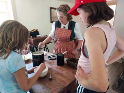 Two young girls observing and working at a table with a period-costumed person in apron.