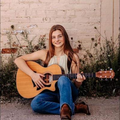 Woman sitting on ground with wall behind; she's holding a guitar.