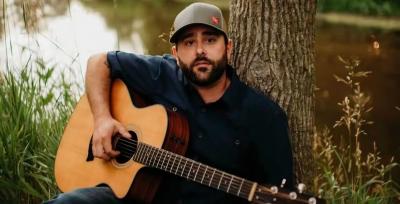 Man in ballcap sitting against tree and holding guitar.