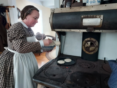 Woman in period dress cooking pancakes on an iron stove.
