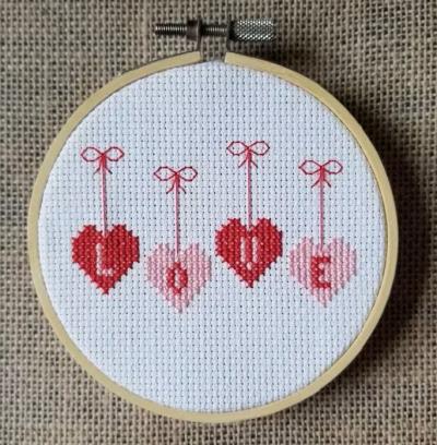 Cross stitch piece in a hoop; four hearts containing letters that spell "LOVE."