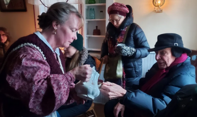Woman in period dress serving wassail to a seated gentleman in winter garb.