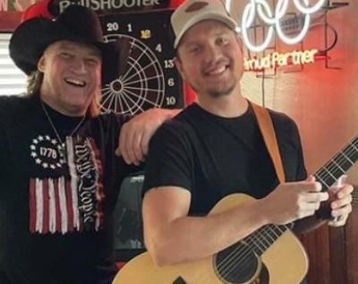 Two smiling men, one in black hat, other in ballcap and holding guitar.