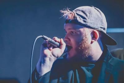 Bearded man in backward ballcap singing into a microphone onstage.