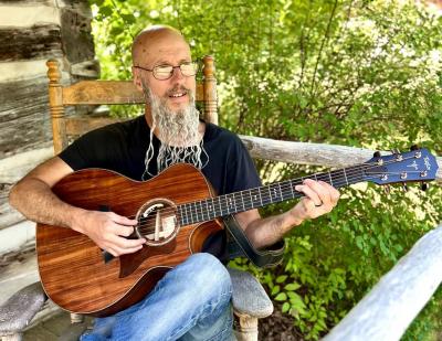 Bearded man seated on a porch and strumming guitar.