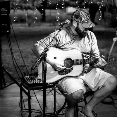B/W photo of man in ballcap sitting outdoors and strumming guitar.