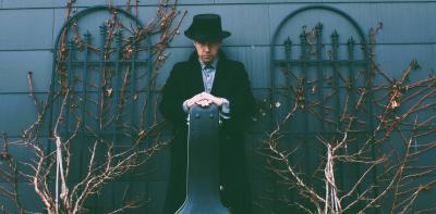 A solemn-looking man in black hat and coat standing against a wall with trellises and holding a guitar case.