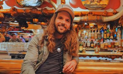 Smiling man with ballcap on seated and leaning back on a bar.