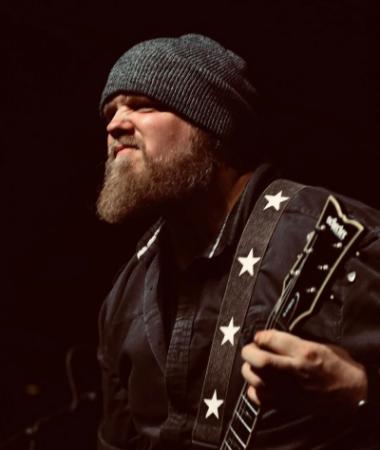 Intense-looking bearded man in a beanie playing guitar onstage.