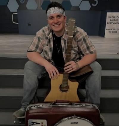 Smiling man with bandana headband seated on steps and holding guitar.