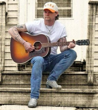 Man in ballcap seated outdoors on some steps and playing guitar.