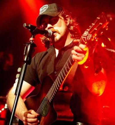 Man in ballcap playing guitar and singing into mike under a red-hued spotlight.