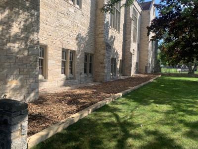 Outer wall of building, an unplanted flower bed and green grass.