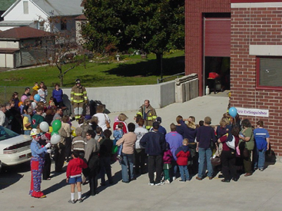 Group of people standing around two firefighters in front of a fire station. Clown in the back of the crowd holding balloons.