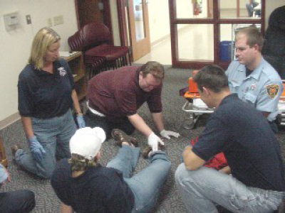 Person lies on the ground. A man wearing white gloves touches their ankle. Three paramedics kneel next to them 