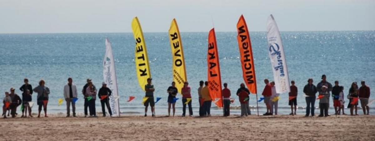 People standing on the beach in front of the ocean. Flags that read Kites Over Lake Michigan