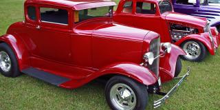 Red classic car next to another on a grassy field.