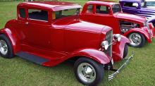 Red classic car next to another on a grassy field.