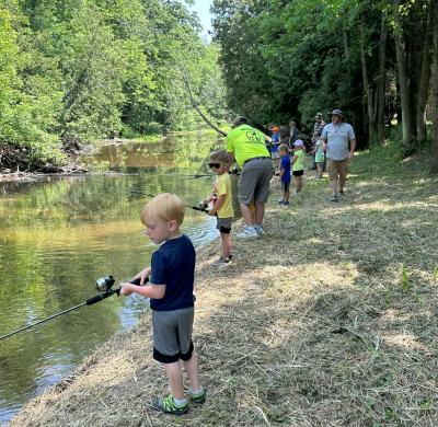 Kids fishing from a riverbank with adults instructing.